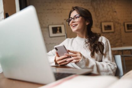 A woman working from a home office, sitting at a desk, holding a phone, and working on a laptop.