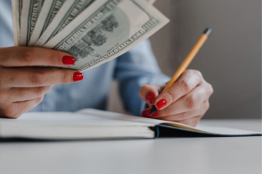 A woman's hands holding and counting cash, while writing in a notebook.