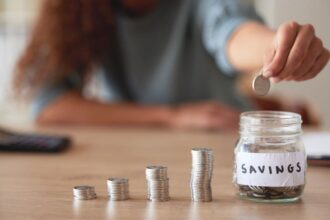 A woman counting coins in piles on a desk and then putting them into a jar labeled "savings".