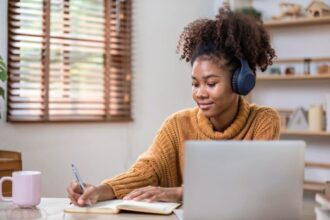 College student working from home, using a laptop, wearing headphones, and writing in a notebook.